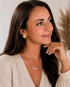 A woman with long dark brown hair is shown in a close-up portrait wearing a gold and pearl jewelry set, including a flower-shaped stud earring and a delicate necklace with a pearl cluster.