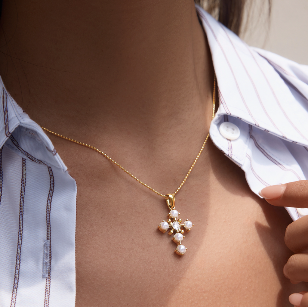 A close-up lifestyle shot of a person wearing a delicate gold cross pendant necklace adorned with white pearls, styled over a white button-down shirt.